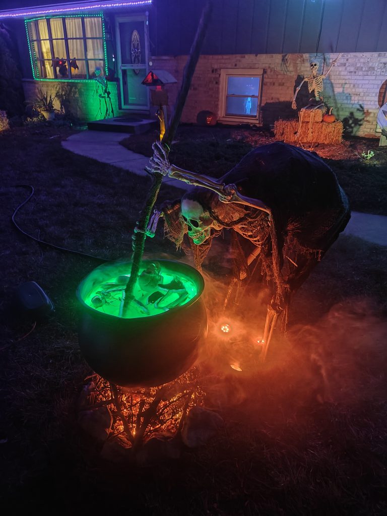 A skeleton wearing tattered fabric stirring a large cauldron that is sitting on a pile of "burning" embers