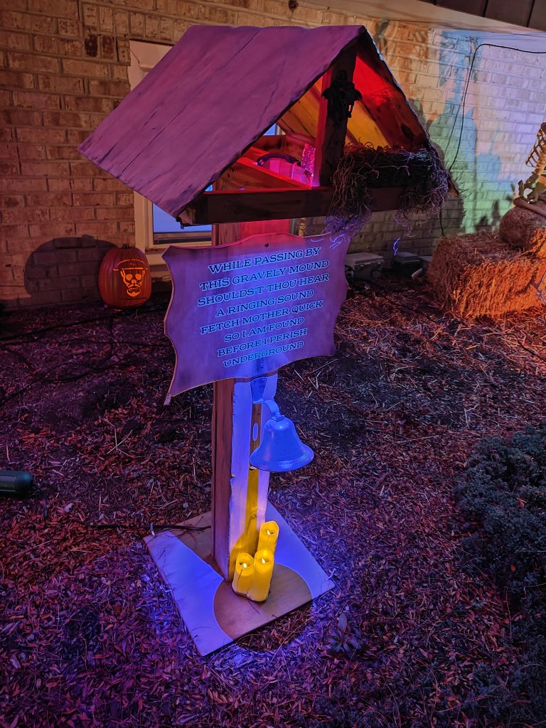 A covered sign post with a bell. The sign reads "When passing by this gravely mound, shoulds't thou hear a ringing sound, fetch mother quick so I am found, before I perish underground"