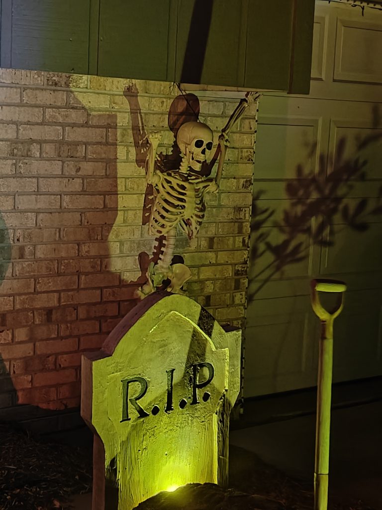 A tombstone with a shovel in front of it and a skeleton behind it.